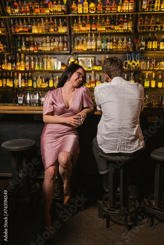 Joyful woman in pink dress flirts with man at pub counter. Pretty lady and guy drink alcohol and communicate in nightclub