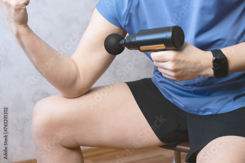 Woman massages the muscles of her arm with a percussion massage gun. Woman massaging hand by handheld massage gun, post-workout recovery routines