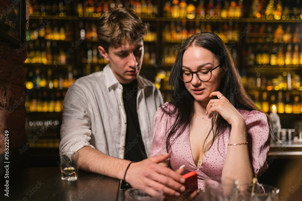Brunette woman with loose hair in silk dress enjoys holiday near friend at blurry wall in pub. Lady in pink dress sits near young man in club