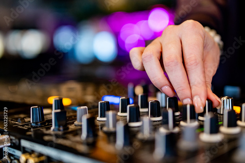 close-up view of a DJ’s hand adjusting knobs on a sound mixer. Various knobs and sliders are visible, indicating different channels and controls for audio mixing. The DJ’s hand is in focus
