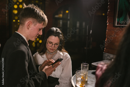 Man with phone tells story to girlfriend near bar table in pub. Young guests communicate and have fun in fancy night club party