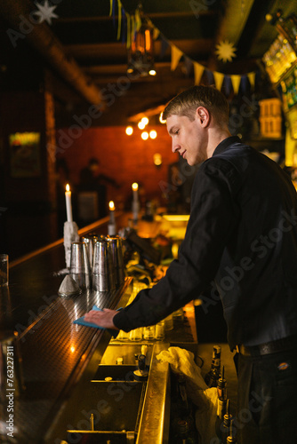 Grinning bartender prepares bar counter for working evening cleaning with rag. Young worker waits for customers in vintage pub