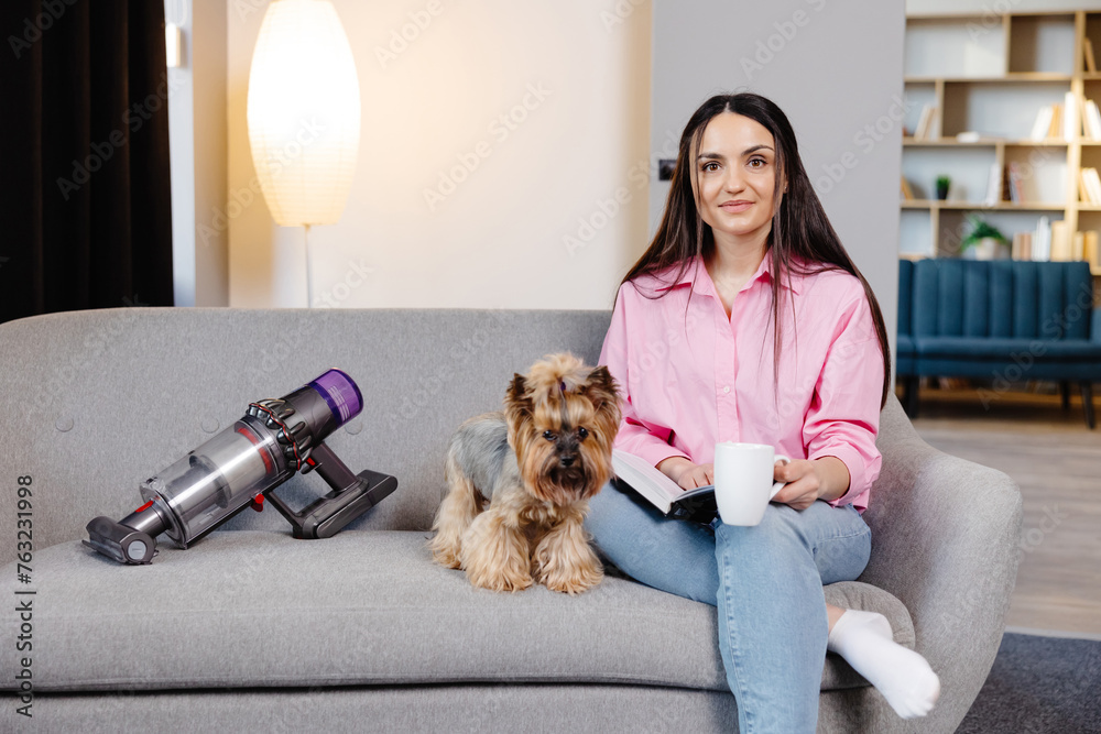 A cute young woman is sitting on the sofa with her dog and reading a book and holding a cup in her hand. A cute young woman is sitting on the sofa with her dog and reading a book and holding a cup in her hand.