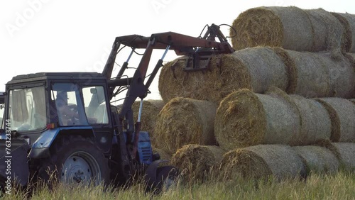 The tractor uses a hydraulic manipulator to stack round bales of hay in pyramids for storage and drying. Machinery works in the field during harvesting.