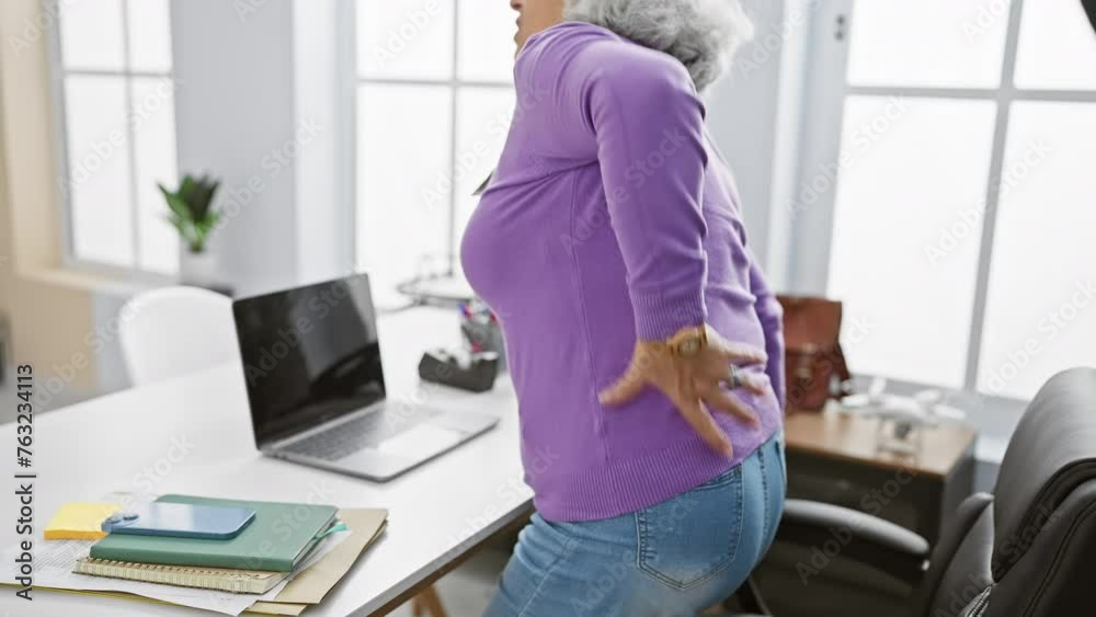 Mature woman in purple experiencing back pain at her clean, well-lit office environment, near a laptop and notepads. Mature woman in purple experiencing back pain at her clean, well-lit office environment, near a laptop and notepads.