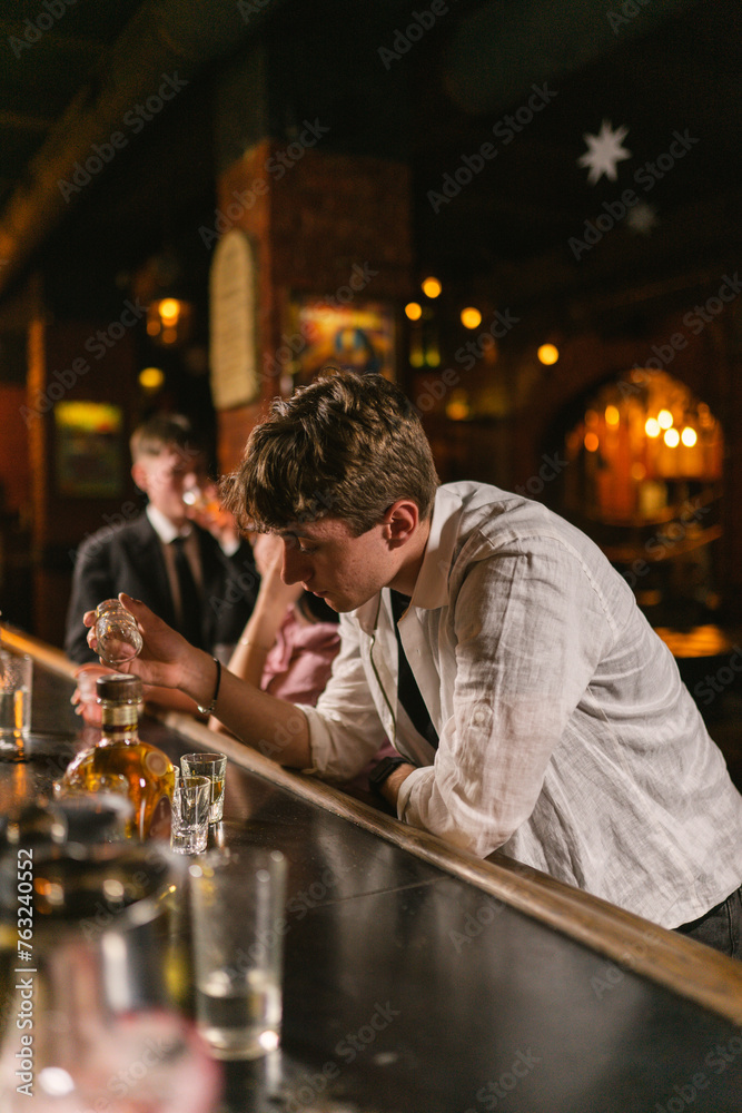 Young man drinks various alcohol alone and thinks about work in cozy pub. Young guy thinks about problems with glass of cognac on bar