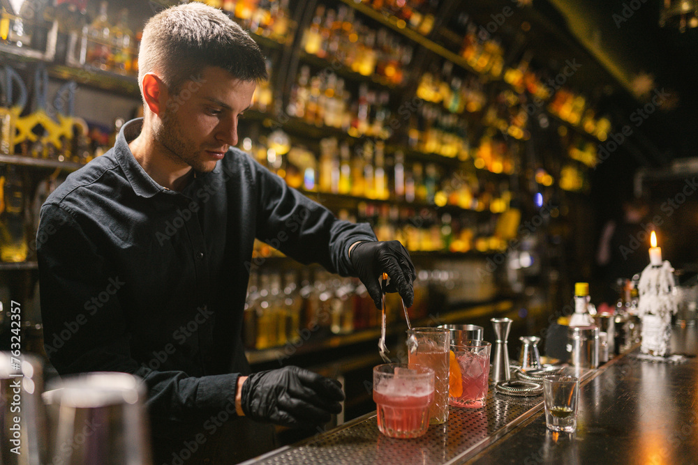Barkeeper in uniform adds ice to glass to cool beverages. Skilled worker of vintage elite bar cooks expensive cocktails for guests