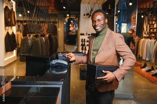 Happy african shopper holding boxes of new clothes or shoes and paying with phone photo