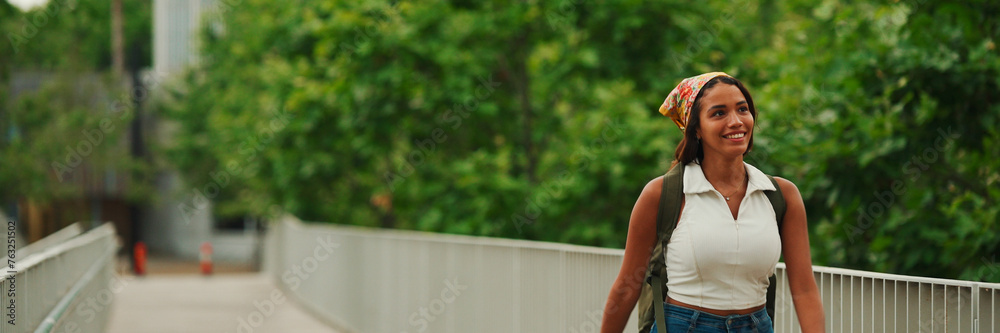 Cute tanned woman with long brown hair in white top and yellow bandana with backpack on her shoulders walks along the bridge, Panorama