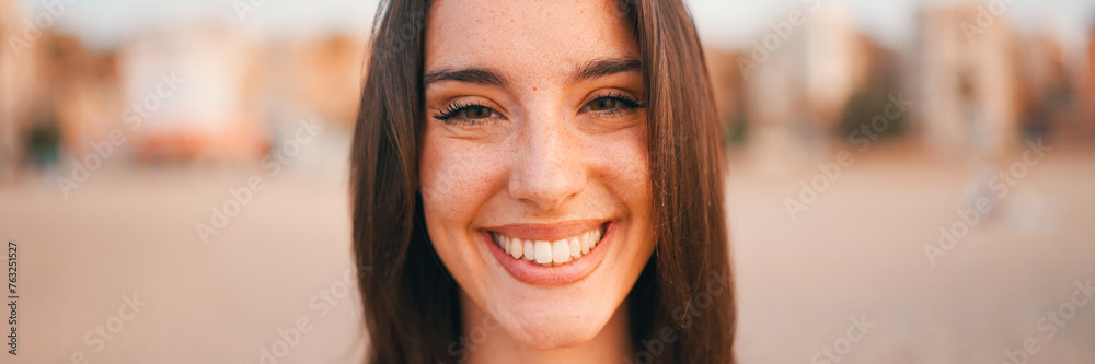 Fototapeta premium Beautiful brown-haired woman with long hair sits on the beach, Panorama. Gorgeous girl with freckles and long eyelashes looks at the camera and smilig.