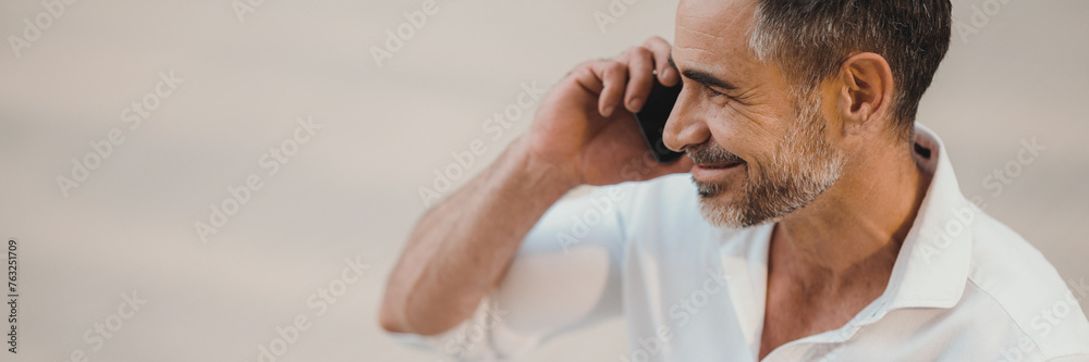 Obraz premium Close up mature businessman with neat beard uses mobile phone sits on bench in the financial district in the city, Panorama. Successful man talking on smartphone.