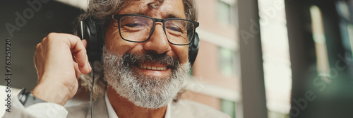 Panorama, Close up portrait of mature businessman with beard in eyeglasses and headphones, sits in an outdoor cafe and works using tablet