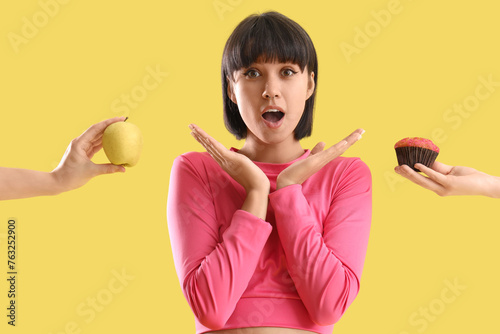 Shocked sporty young woman receiving apple and muffin on yellow background. Diet concept