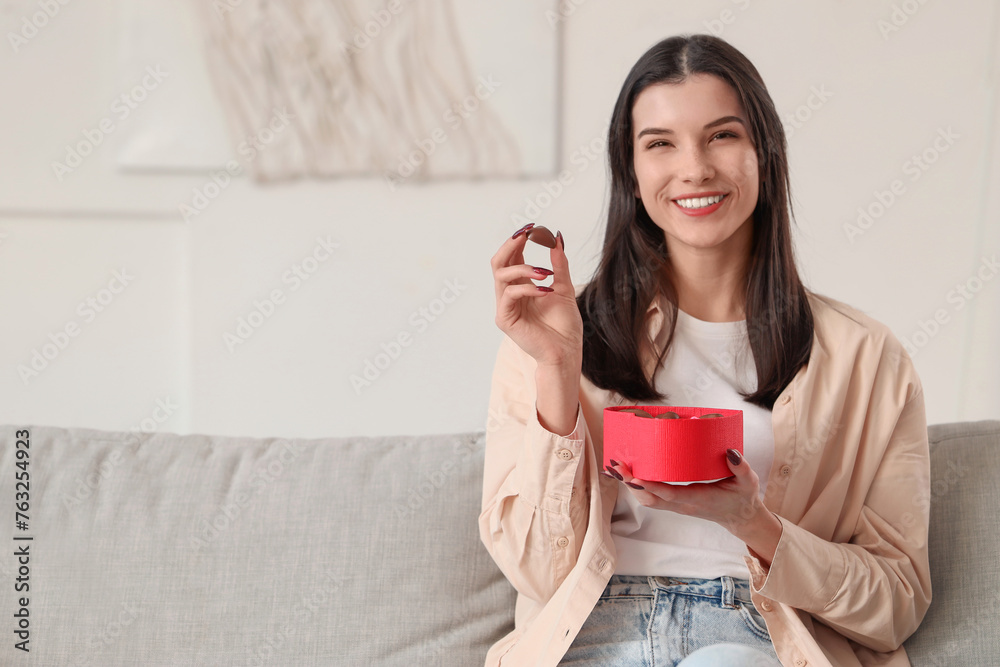 Young woman with box of heart-shaped chocolate candies sitting on sofa at home Young woman with box of heart-shaped chocolate candies sitting on sofa at home