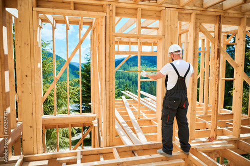 Carpenter constructing wooden-framed dwelling. Back view of man measures distances using tape measure, dressed in workwear and helmet. Idea behind modern, environmentally-conscious building practices.