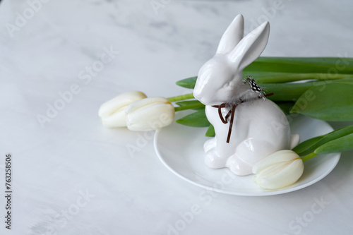 Aesthetic minimal Easter holiday decoration, white porcelain bunny figurine on plate, tulip flowers on neutral marble table background with soft natural light, copy space