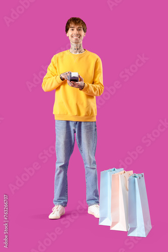 Young online store seller with payment terminal and shopping bags on purple background