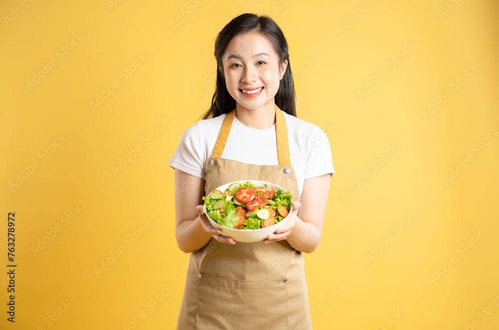 Portrait of Asian female housewife holding a bowl of salad and posing against a yellow background