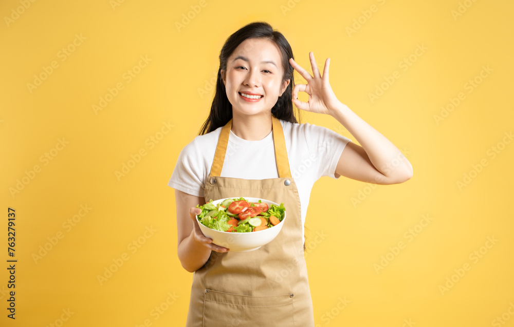 Portrait of Asian female housewife holding a bowl of salad and posing against a yellow background