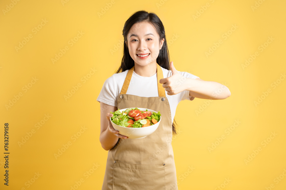 Portrait of Asian female housewife holding a bowl of salad and posing against a yellow background Portrait of Asian female housewife holding a bowl of salad and posing against a yellow background