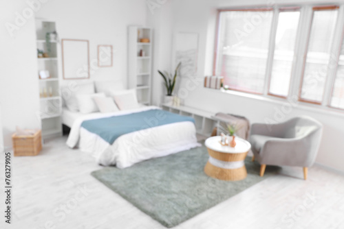Blurred view of light bedroom with big bed  shelf units and frames