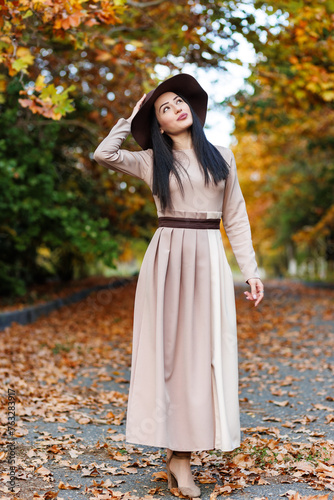 A stylish young woman in fashionable attire standing in autumn park and touching her wide-brimmed hat