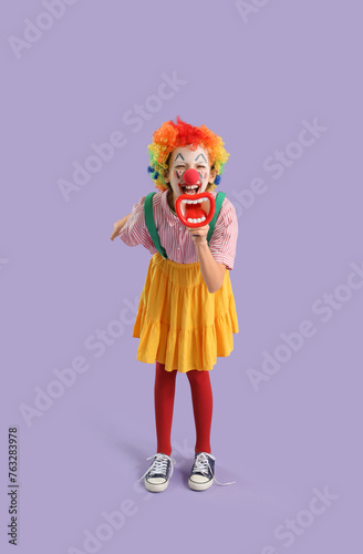 Funny little girl dressed as clown shouting into megaphone on lilac background. April Fools  Day celebration