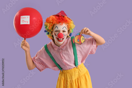 Funny little girl dressed as clown with balloon and paper fish on lilac background. April Fools' Day celebration