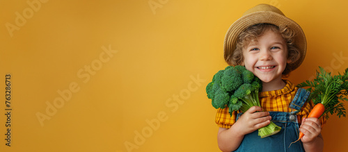A studio shot of a smiling Caucasian boy in a straw hat holding fresh broccoli and carrots on a yellow background. Copying the space. The concept of healthy baby food.