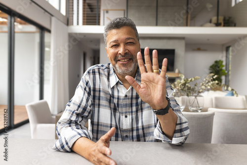 A middle-aged biracial man is waving at the camera in a video at home