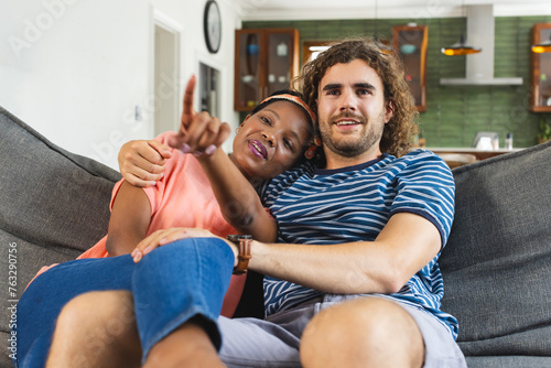 A diverse couple relaxes on sofa at home, pointing at something while watching TV