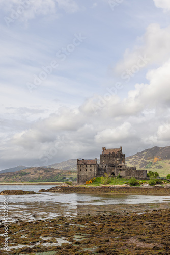 Eilean Donan Castle, poised on the edge of a loch, showcases the rugged beauty and heritage of the Highlands, with a serene sky and low tide revealing the algae-covered shore (Vertical photo)