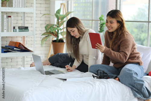 Two young Asian female friends, roommates, sitting on the bed, doing homework together, using laptops and making reports to send to the university, helping each other stay in the apartment. photo