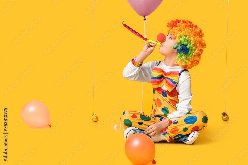 Funny little boy in clown costume with party whistle and balloons sitting on yellow background. April Fools Day celebration - obrazy, fototapety, plakaty 