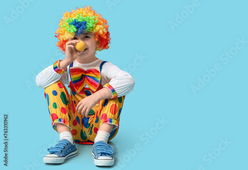 Funny little boy in clown costume sitting on blue background. April Fools Day celebration