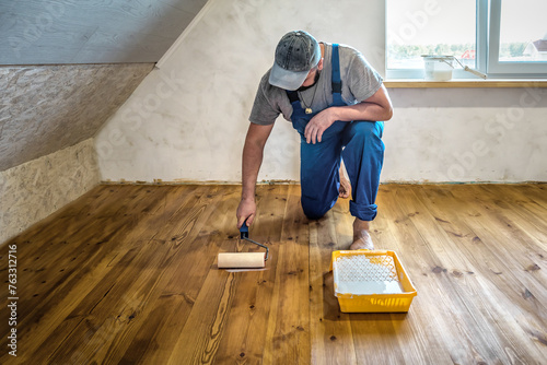 Worker in blue overalls laquering the wooden floor with paint roller and eco varnish