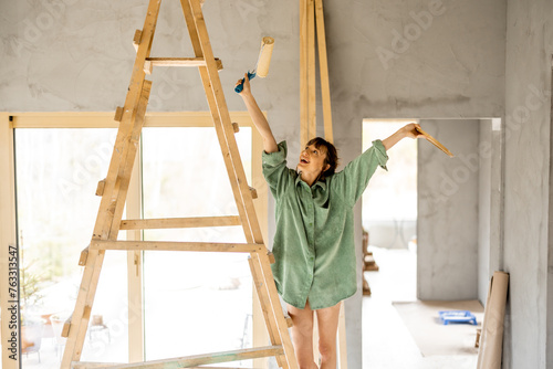 Portrait of a young cute woman standing happily on a ladder with paint roller during repairing process of a house. Creative process of home renovation and repair concept
