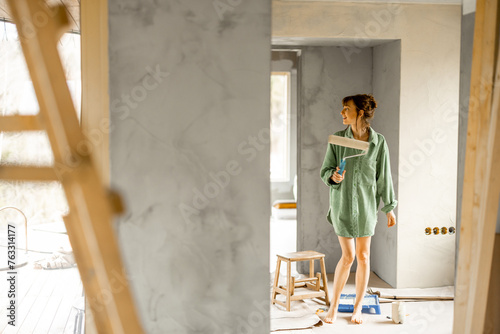 Portrait of a young cute woman standing with paint roller and bucket full of paint during repairing process of a house. Creative process of home renovation and repair concept