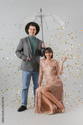 Elegant young couple with umbrella and falling confetti on light background