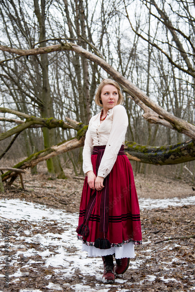 romantic single  beautiful blonde girl in ukrainian traditional clothes and red skirt and boots staying near tree in empty winter field with snow