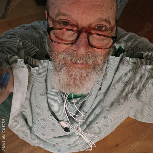 Man Senior, in a hospital robe, looking up at the camera. Wearing a heart monitor device. Beard and glasses.
