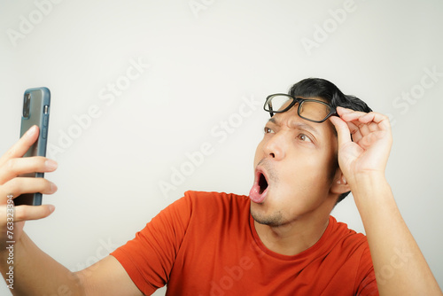 Wow face of Asian man in orange t-shirt shocked what he see in the smartphone on isolated background