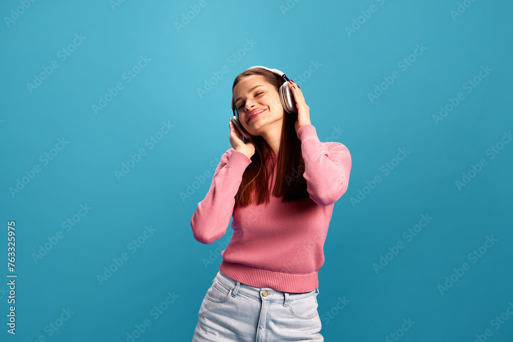 Smiling and positive young woman in pink sweater and jeans dancing and listening to music in headphones against blue studio background. Concept of youth, lifestyle, casual fashion, human emotions Smiling and positive young woman in pink sweater and jeans dancing and listening to music in headphones against blue studio background. Concept of youth, lifestyle, casual fashion, human emotions