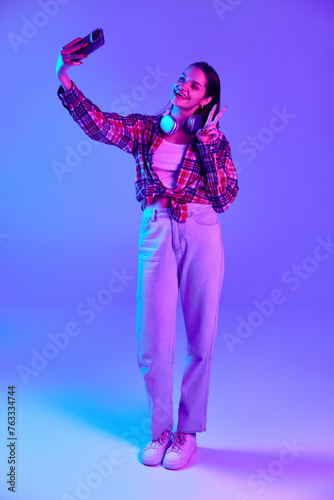 Full-length image of smiling young woman in checkered shirt taking selfie with mobile phone against purple studio background in neon light. Concept of youth  lifestyle  casual fashion  human emotions