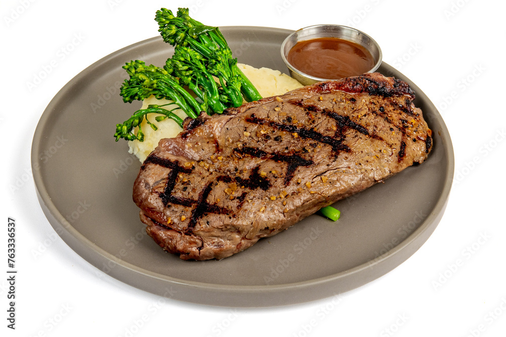 A strip loin steak showing grill marks on a plate with broccolini and mashed potatoes and a ramekin of gravy isolated on white
