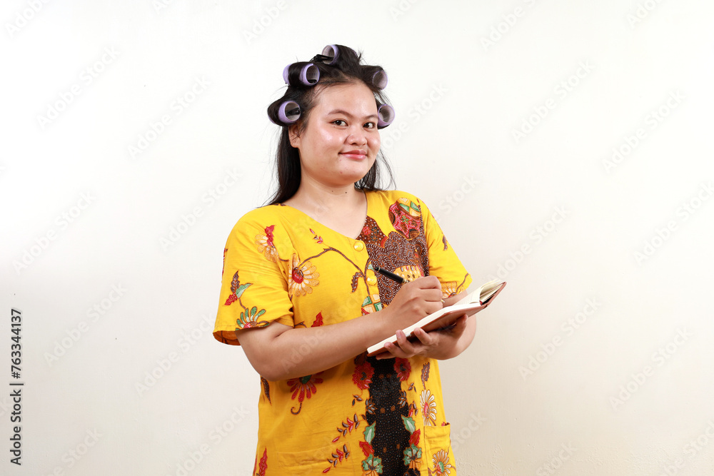 Joyful asian housewife standing while writing on the book and looking at the camera Joyful asian housewife standing while writing on the book and looking at the camera