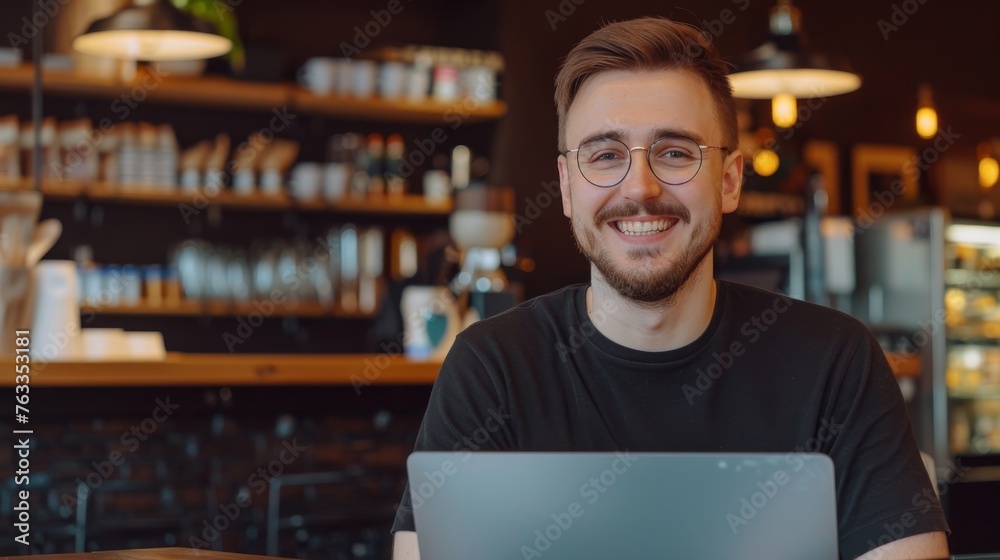 A happy young man with a laptop, sitting in a coffee shop, the portrait image showcasing a modern professional working remotely