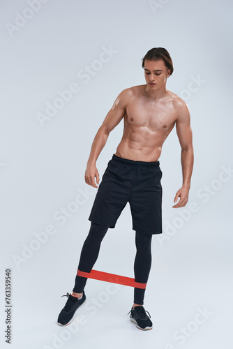 alluring sporty young man in black pants posing topless with resistance band and looking away