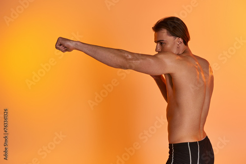 alluring shirtless sexy man in black sporty shorts boxing actively on vibrant orange background
