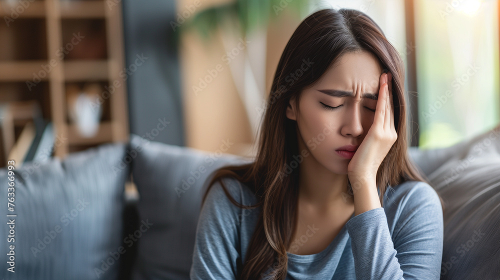 Dizzy asian young woman, girl headache or migraine pain suffering from vertigo while sitting on couch in living room at home, holding nose with hand, health problem of brain or inner ear not balance.